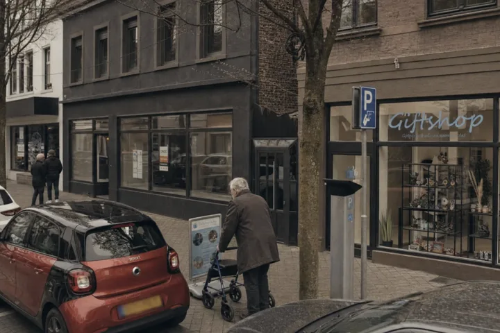 Vrouw met rollator moet tussen een reclamebord en een geparkeerde auto door