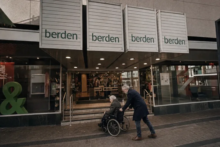 Man duwt een rolstoelgebruiker naar een winkel waar alleen een trap naar de ingang leidt