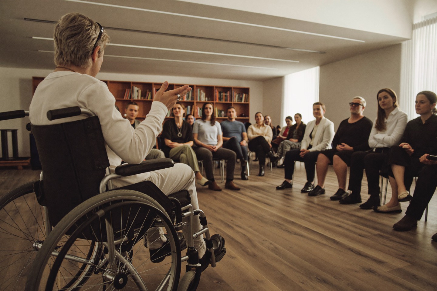 Een spreker in een rolstoel spreekt een aandachtig publiek toe in een lichte zaal